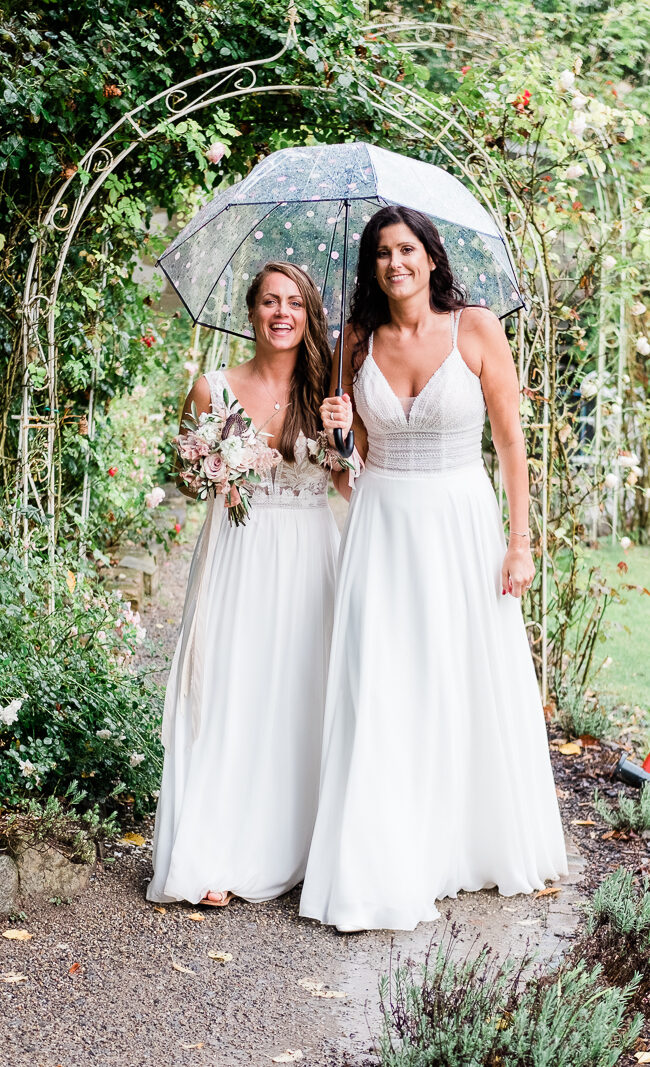 Zwei Bräute in weißem Kleid unter einem Regenschirm im Rosengarten
